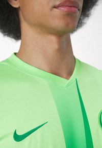 La maglia Nike verde presenta un design a V, una texture liscia e un logo swoosh verde scuro sul davanti, con sottili strisce tonali.