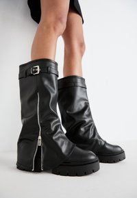 Las botas de cuero negras hasta la rodilla cuentan con un eje ancho con una hebilla ajustable y una cremallera lateral, complementadas por una suela gruesa.