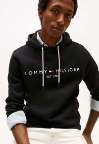 Sudadera negra con cordones blancos, con texto y logo "TOMMY HILFIGER" en rojo. Lleva puesta sobre una camisa de rayas con cuello y puños arremangados.