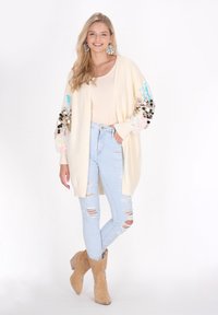 Cremefarbener Cardigan mit Paillettenakzenten an den Ärmeln, kombiniert mit hellblauen, zerrissenen Jeans und sandfarbenen Ankle-Boots. Weicher, texturierter Stoff.