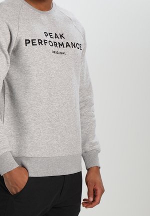 Osoba ubrana w jasno szarą bluzę z napisem „PEAK PERFORMANCE ORIGINAL”, ręce rozluźnione, na jednolitym jasnym tle.