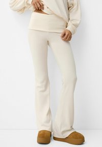 Cream flared pants in soft fabric with ribbed waistband. Paired with tan, suede-like slip-on shoes. Clean lines and minimal design.