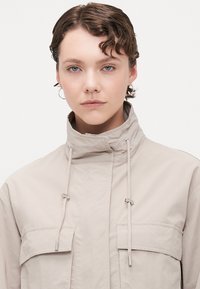 Light beige jacket with a high collar, drawstring closure, and two chest pockets. Smooth fabric with a matte texture.