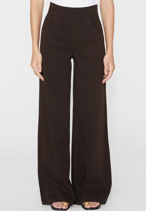 Donna che indossa pantaloni neri a vita alta e gamba larga con tasche frontali e sandali neri con anelli d'oro alle dita, in piedi su uno sfondo bianco.