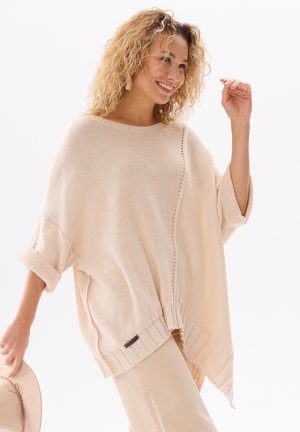 Lächelnde Frau mit lockigem Haar trägt einen übergroßen beigen Strickpullover und helle Hosen, hält einen beigen Hut in einer Hand vor einem weißen Hintergrund.