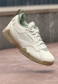 Zapatilla deportiva en color beige claro y verde con cordones, suela texturizada, panel lateral de malla y un logo que se asemeja a una figura saltando sobre una superficie de hormigón.