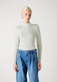 Skaidri zaļš garu piedurkņu augstais apkakles tops, izgatavots no gludas auduma, apvienots ar augsto jostasvietu zilām denim biksēm, kas ir brīvas piegriezuma un ar krokām.