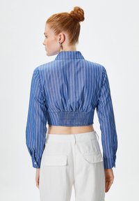 Camicia blu a righe cropped con maniche lunghe, dotata di un retro arricciato e colletto. Abbinata a pantaloni bianchi, vista posteriore.