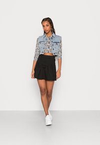 Colete de ganga com fecho à frente, blusa de manga longa com padrão de zebra e cropped, e saia mini preta plissada. Modelo usa ténis brancos.