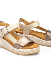 Sandalias de cuña con tiras de cuero dorado y crema, hebilla ajustable y suela de plataforma blanca texturizada. Plantilla suave color marrón claro. Forma redondeada.
