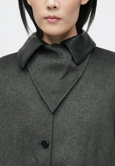 Pilkas vilnonis paltas, pasižymintis unikaliu juodos odos smailiu apykakliu, su vienu juodu mygtuku ir glotnia, tekstūrine danga.