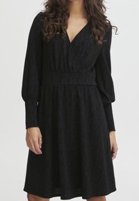 Czarna sukienka z długimi rękawami, o kroju kopertowym, z teksturowanej tkaniny i dopasowanej talii. Rozkloszowana spódnica sięga tuż ponad kolano.