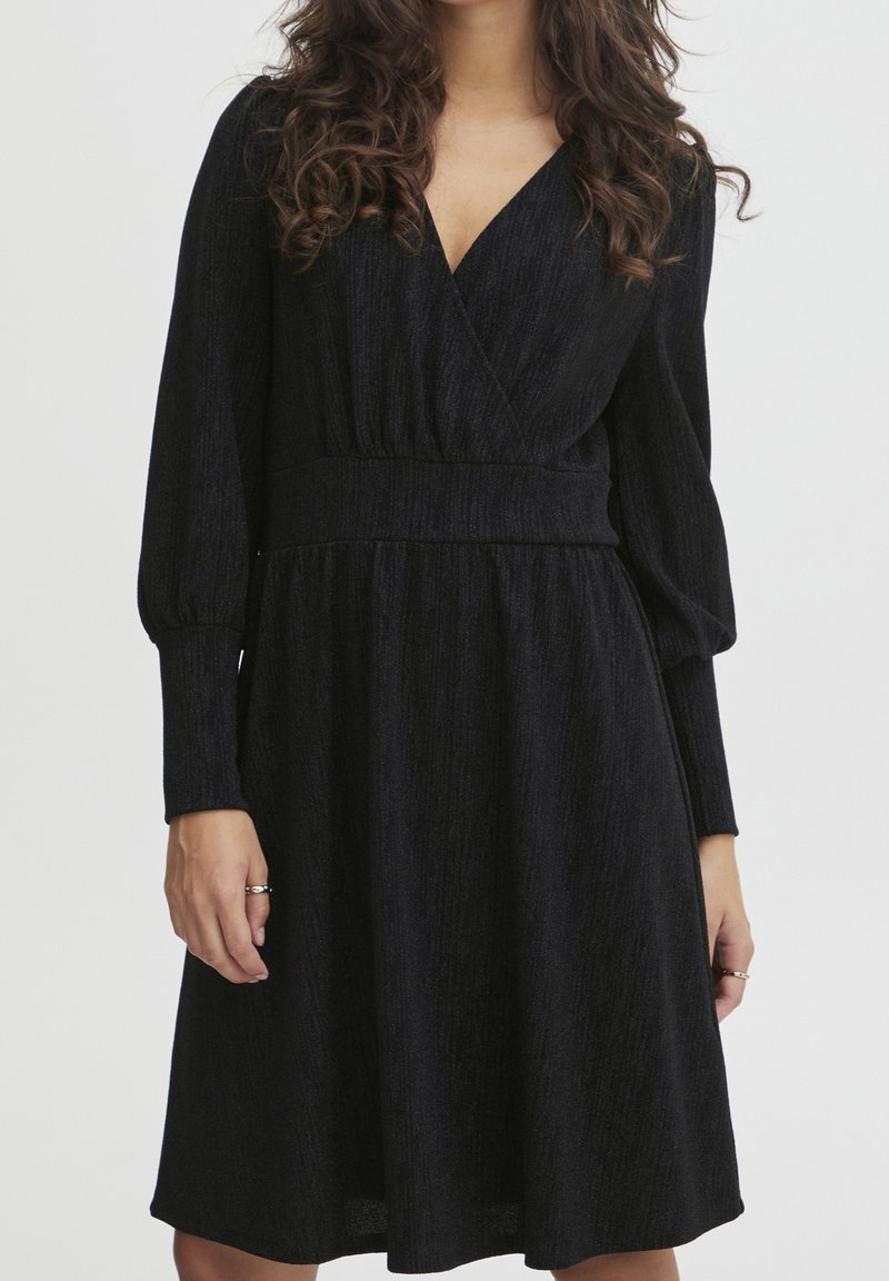 Czarna sukienka z długimi rękawami, o kroju kopertowym, z teksturowanej tkaniny i dopasowanej talii. Rozkloszowana spódnica sięga tuż ponad kolano.