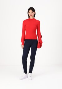 Crvena rebrasta džemper s dugim zvonastim rukavima, uparen s tamnoplave slim-fit hlačama i bijelim tenisicama. U jednoj ruci drži malu crvenu torbicu preko ramena.