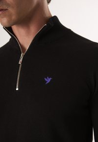 Černý pletený svetr s vysokým límcem, který má stříbrný zip a malé fialové ptáčkové logo na hrudi.