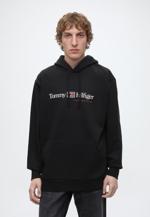 Млад мъж със светла къдрава коса носи черен суитшърт Tommy Hilfiger и тъмни дънки, стоящ на бял фон.