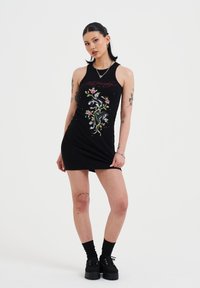 Schwarzes ärmelloses Kleid mit floraler Stickerei und Text, figurbetontes Design, gerippte Textur, kombiniert mit schwarzen Plateauschuhen und Socken.