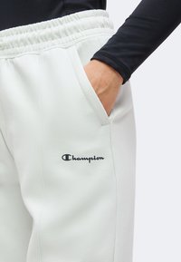 Χέρι στην τσέπη λευκών φούτερ παντελονιών Champion, συνδυασμένα με μαύρο μακρυμάνικο τοπ, δείχνοντας ελαστική ζώνη και λογότυπο στο μηρό.