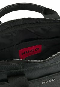 Zwarte leren tas met een gestructureerde afwerking, voorzien van een ritssluiting en een rood merklabel binnenin, glad zwarte voering en dubbele handvaten.
