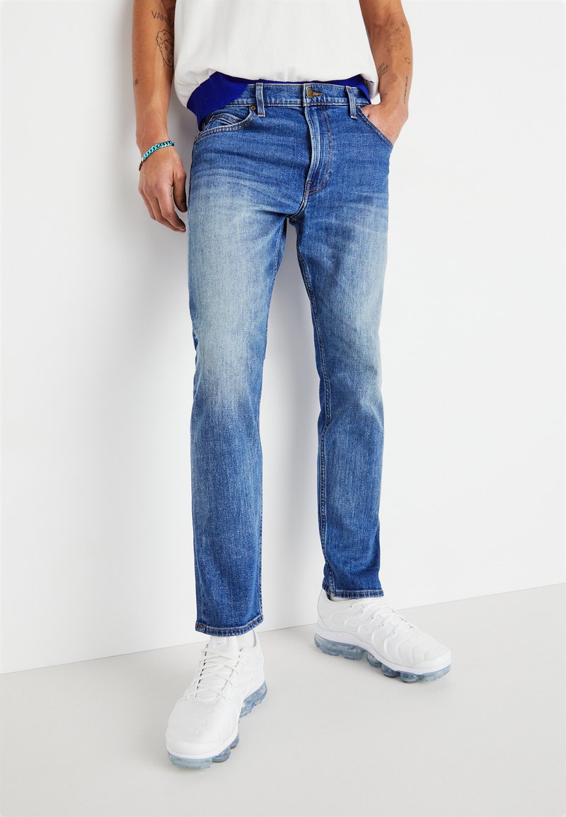Light blue denim jeans with a slim fit, featuring a faded finish, five pockets, and a button closure, paired with white athletic shoes.