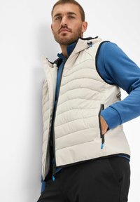 Gilet trapuntato beige chiaro con cerniera e due tasche laterali, con dettagli blu e cappuccio. Indossato sopra una maglietta a maniche lunghe blu.
