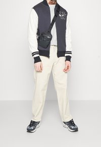 Marineblauw-witte bomberjack met ritssluiting aan de voorkant en logo, grijs T-shirt, crème broek en zwarte sneakers met grijze accenten. Schoudertas bevestigd.