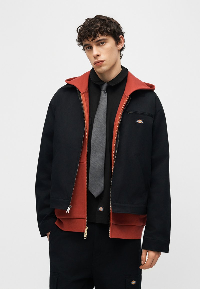 Junger Mann mit lockigem Haar, der eine schwarze Jacke über einem rostfarbenen Hoodie, einem schwarzen Hemd, einer grauen Krawatte und schwarzen Hosen vor einem schlichten Hintergrund trägt.