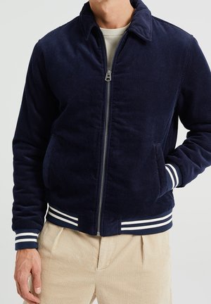 Mann trägt eine marineblaue Cordjacke mit Reißverschluss, weißen gestreiften Bündchen und Saum, beigefarbene Cordhose, eine Hand in der Jackentasche.