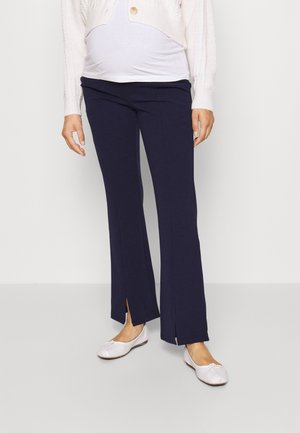 Pantaloni premaman blu navy con spacco alla caviglia, realizzati in morbido tessuto. Abbinati a una leggera maglietta bianca e a delle scarpe basse di colore chiaro.
