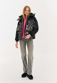 Młoda kobieta uśmiechająca się, ubrana w czarną pikowaną kurtkę z kwiatowym wzorem, różowy sweter, szare dżinsy i czarne buty na platformie, stojąca na białym tle.