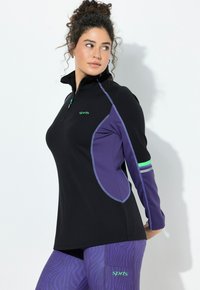 Žena s kudrnatými vlasy, která má na sobě černý a fialový sportovní top a fialové legíny s vzorem, hledící stranou na bílém pozadí.