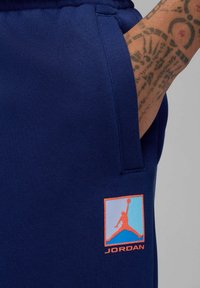 Hånd med tatovering i lommen på marineblå træningsbukser med et multifarvet Jordan-logo-patch nær låret.