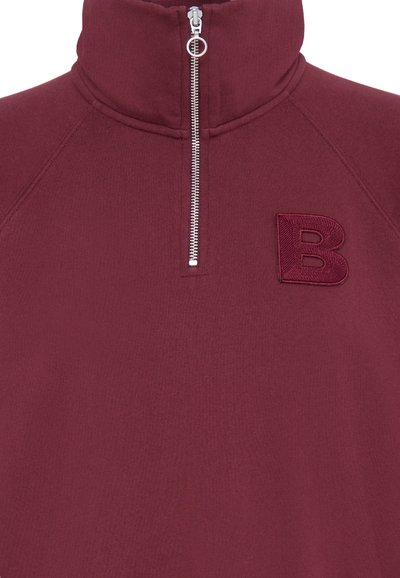 Burgundska zip-up majica s visokom ovratnikom, s srebrnim cipkom i izvezenim slovom 'B' na prsima. Mekan materijal, rebrasti detalji.