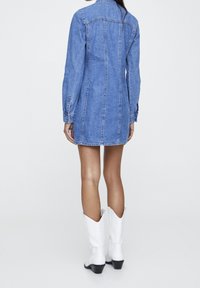 Robe chemise en denim à manches longues, couleur bleu clair, avec coutures latérales. Portée avec des bottes de cowboy blanches ornées de coutures décoratives.