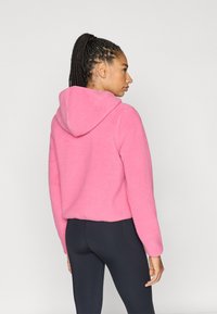 Różowa bluza z kapturem z polaru o dopasowanym kroju, kapturem i długimi rękawami. Tkanina wydaje się miękka i teksturowana, z krótkim krojem sięgającym talii.