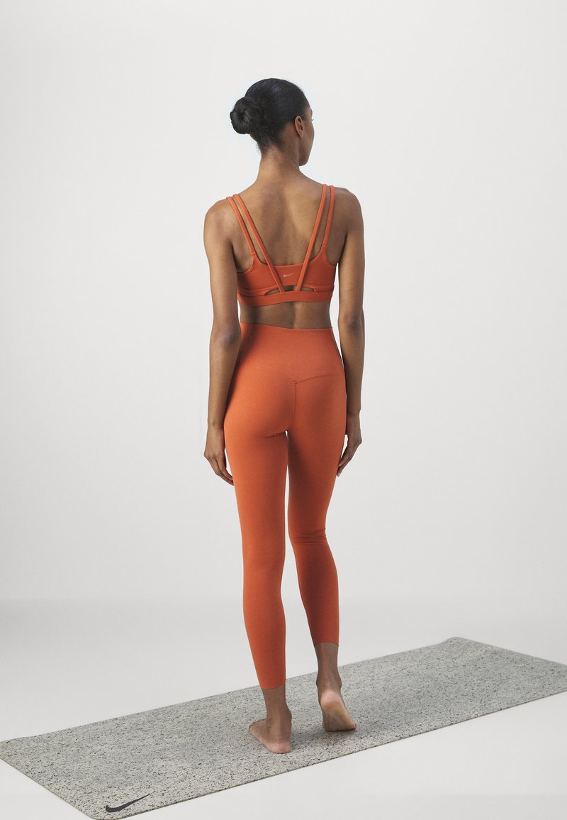 Reggiseno sportivo arancione con più cinghie, abbinato a leggings coordinati. Il modello è in piedi a piedi nudi su un tappetino da yoga grigio.