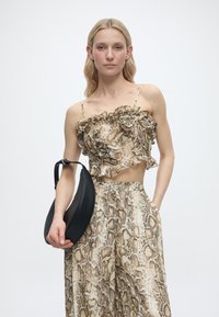 Γυναίκα που φοράει κορυφή με βολάν και τύπωμα φιδιού και ταιριαστό παντελόνι με φαρδιές παντελόνες, κρατώντας μια μαύρη τσάντα σε σχήμα ημισελήνου με ρυθμιζόμενο λουρί.