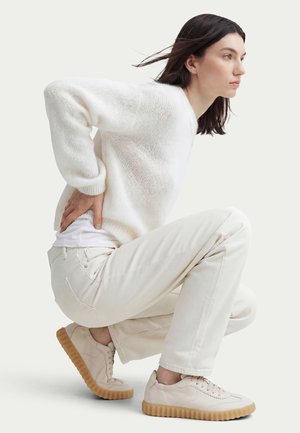 Donna con maglione bianco, pantaloni beige e sneakers beige con suole in gomma accucciata, che si tiene la parte bassa della schiena, guardando avanti su uno sfondo uniforme.