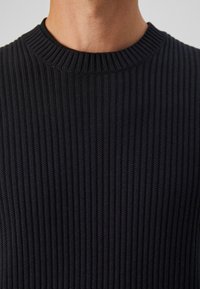 Close-up of a person wearing a black ribbed knit sweater with a crew neckline, showing the neck and upper chest area.