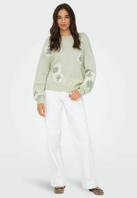Pull vert clair avec des accents floraux, encolure ronde et épaules tombantes, associé à un pantalon large blanc et des ballerines marron.