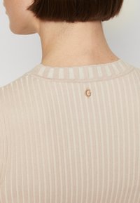 Haut côtelé beige clair avec un col rond et des rayures verticales. Présente un petit accent de logo métallique à l'arrière. Texture douce.