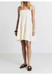 Robe blanche à étages avec fines bretelles et tissu texturé, dotée d'un corsage ajusté et d'un ourlet à volants, associée à des chaussures tissées foncées.