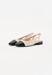 Chaussures à talon ouvert beiges et noires avec un bout pointu, présentant un capuchon noir brillant et un motif découpé sur les côtés. Talon plat.