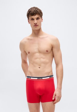 Νέος άνδρας με κοντά καστανά μαλλιά που φοράει κόκκινα ανδρικά εσώρουχα Levi's boxer briefs, στέκεται μπροστά σε ένα απλό ανοιχτό γκρι φόντο.