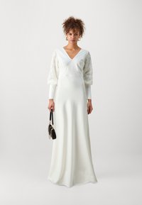 Witte satijnen lange jurk met een V-hals, pofkanten mouwen en een vloeiende silhoutte, gecombineerd met een kleine zwarte handtas.