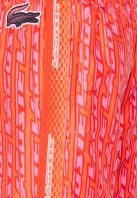 Shorts de bain orange avec un motif texturé rose, doté d'un logo noir et d'une inscription blanche sur le côté. Ceinture élastique incluse.