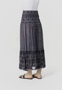 Magasderekú, bokáig érő szoknya tengerészkék és szürke geometriai mintákkal, csupasz lábon fekete szegecses lapos szandállal viselve.