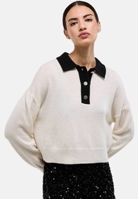 Pull crème avec un col noir et des boutons ; caractérisé par une coupe décontractée et des manches longues, associé à une jupe noire à sequins.
