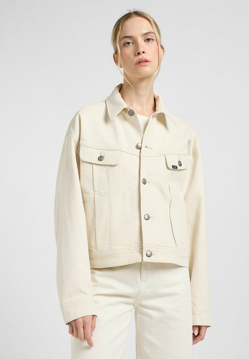 Veste en denim beige clair avec un col, des boutons-pression, des poches poitrine et une coupe décontractée. Texture lisse avec des détails de couture visibles.