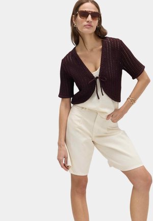 Kvinde iført mørkebrun kortærmet strikket cardigan bundet ved brystet, hvid ærmeløs top, cremefarvede Bermuda-shorts, solbriller og guldarmbånd.